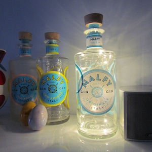 May include: Three clear glass bottles of Malfy Gin with blue and yellow labels, and cork stoppers. One bottle is lit with fairy lights. The bottles are on a white surface, next to a speaker and a polka-dot pig figurine.