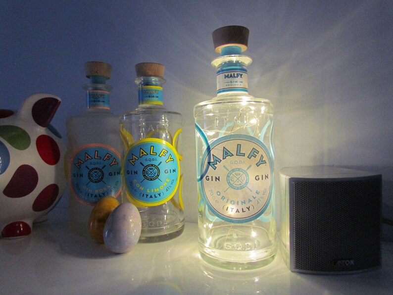 May include: Three clear glass bottles of Malfy Gin with blue and yellow labels, and cork stoppers. One bottle is lit with fairy lights. The bottles are on a white surface, next to a speaker and a polka-dot pig figurine.