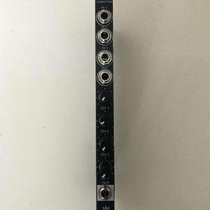 Op de afbeelding: Een zwart elektronisch module met meerdere ingangs- en uitgangscontacten, knoppen en de tekst "COMTENTEN". De module heeft ronde ingangspoorten met de labels IN 1, IN 2, IN 3 en IN 4. Het heeft ook drie kanaalknoppen met de labels CH 1, CH 2 en CH 3.