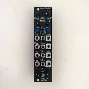 Puede incluir: Módulo de sintetizador modular negro con perillas, conectores y texto. El módulo tiene el texto "QUAD VOLTAGE CONTROLLED AMPLIFIER/VC MIXER MODULE" y "BLK BLK". Cuenta con múltiples entradas y salidas, con el texto "MODULAIRE MARITIME" en la parte inferior.