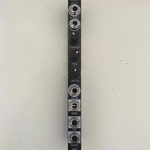 May include: Black electronic module featuring multiple input/output jacks, knobs, and text labels. The module has circular jacks, black knobs, and text including "essaim", "WOCT", "H SYNC", "COARSE", "FINE", "LIN FM", "FM IN", "SINE", "TRI", "SSAW", and "MM".