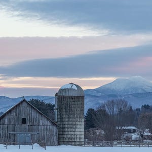 Puede incluir: Un granero de madera desgastado y un silo se alzan en un campo nevado con un fondo de montañas cubiertas de nieve y un cielo azul pálido. El cielo es de un rosa y púrpura suave en el horizonte.