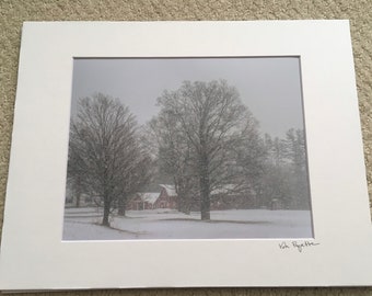 Puede incluir: Una fotografía en blanco y negro de un granero rojo en un campo nevado con dos árboles desnudos en primer plano. Los árboles están cubiertos de nieve y el cielo está nublado. La fotografía está firmada "Kate Royette" en la esquina inferior derecha.