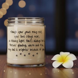 Op de afbeelding: Een glazen potkaars met een witte was binnenkant. De kaars heeft een gedrukte boodschap en pootafdrukken. Rechts van de kaars staat een wit-gele bloem. De achtergrond is vervaagd met bokeh-lichten.