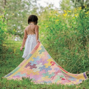 May include: A colorful patchwork quilt with a variety of pastel colors, including pink, yellow, and blue. The quilt is being held up in a grassy field. The quilt has a geometric pattern.