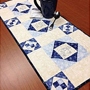 May include: A white and blue quilted table runner with a snowflake design. The runner is decorated with a blue mug and a silver spoon.