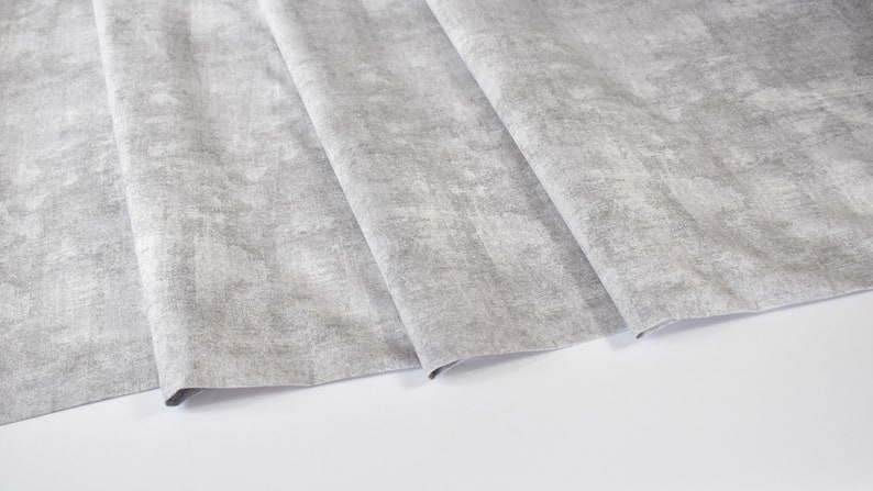 May include: A close-up of a gray fabric with a textured, slightly distressed look. The fabric is folded in several layers, showing the texture and pattern.