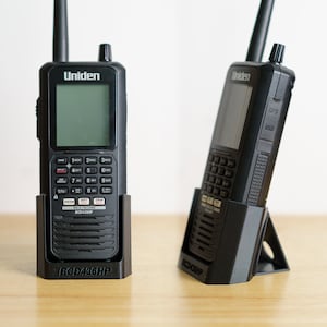 Puede incluir: Dos escáneres Uniden BCD436HP negros con una base de carga negra. Los escáneres tienen una pequeña pantalla y un teclado con botones numerados del 1 al 9, A, B, C, D, E, F, G, H, I, J, K, L, M, N, O, P, Q, R, S, T, U, V, W, X, Y, Z y 0. Los escáneres están en una base de carga con el texto "BCD436HP" impreso en la parte delantera.