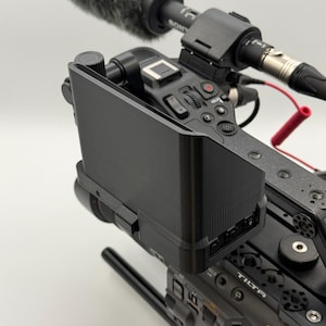 May include: A black matte monitor sun hood attached to a professional video camera. The camera body features a Tilta mounting cage, a top handle with multiple screw holes, and a Sony microphone mounted on top with a red cable connection.