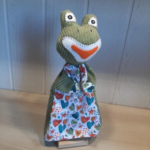May include: A plush frog doll with a green corduroy body, white eyes, and an orange mouth. It wears a scarf and holds a bag with a colorful heart and star pattern. The doll is displayed on a wooden base.