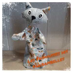 Peut inclure: Marionnette de chat grise avec un motif floral sur le corps et les bras. La marionnette a des yeux noirs et un nez noir. Le texte "LES FRIMOUSSES DES FARIBAMBelles" est écrit en orange en bas de l'image.