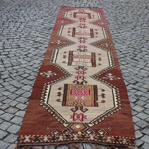 May include: A long, rectangular rug with a traditional Turkish design. The rug is primarily beige with brown, pink, and yellow accents. The design features geometric shapes and intricate patterns.