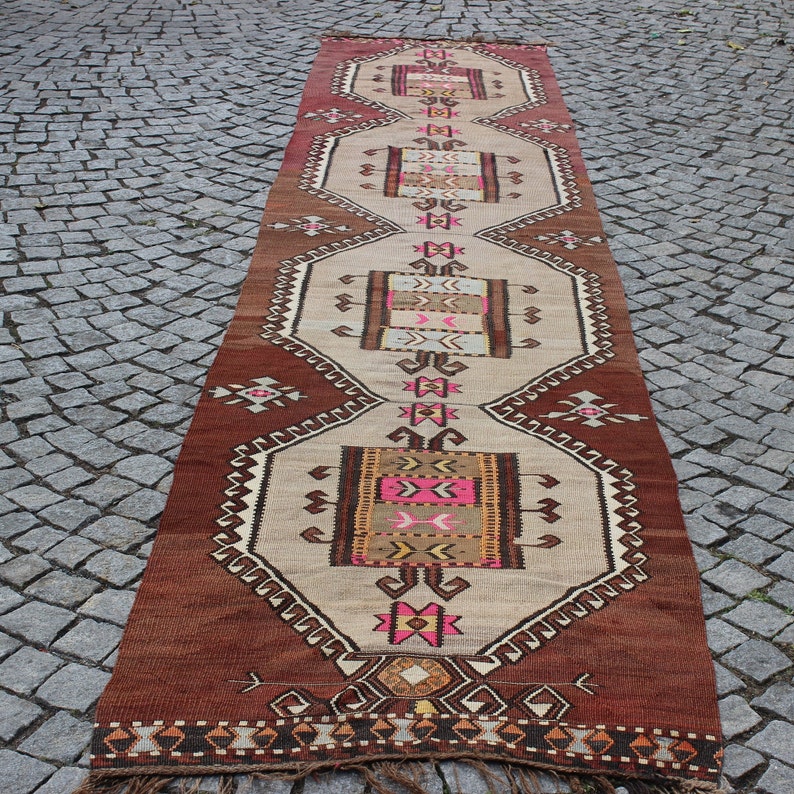 May include: A long, rectangular rug with a traditional Turkish design. The rug is primarily beige with brown, pink, and yellow accents. The design features geometric shapes and intricate patterns.