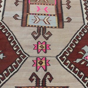 May include: A traditional Turkish rug with a brown and beige background. The rug features a geometric pattern with pink, white, and brown accents. The rug is woven with intricate details and has a fringed edge.