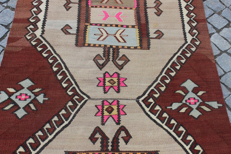 May include: A traditional Turkish rug with a brown and beige background. The rug features a geometric pattern with pink, white, and brown accents. The rug is woven with intricate details and has a fringed edge.