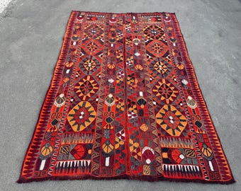 May include: A rectangular textile rug featuring a vibrant red base with intricate geometric patterns in orange, black, and white. The design includes repeating diamond motifs, stylized floral shapes, and zigzag borders with a fringed edge.