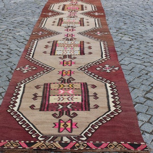 May include: A long, narrow rug with a traditional Turkish design. The rug is beige with red, pink, and black geometric patterns. The rug has a fringe on both ends.