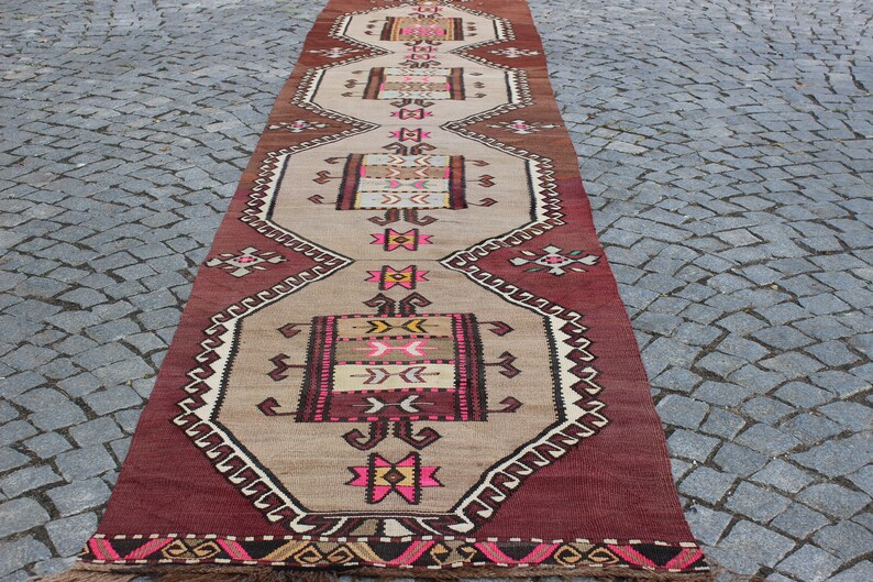 May include: A long, narrow rug with a traditional Turkish design. The rug is beige with red, pink, and black geometric patterns. The rug has a fringe on both ends.