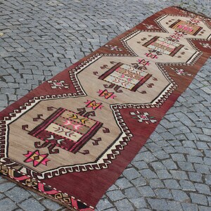 May include: A long, narrow rug with a geometric pattern in shades of red, brown, beige, and pink. The rug has a fringe on both ends.