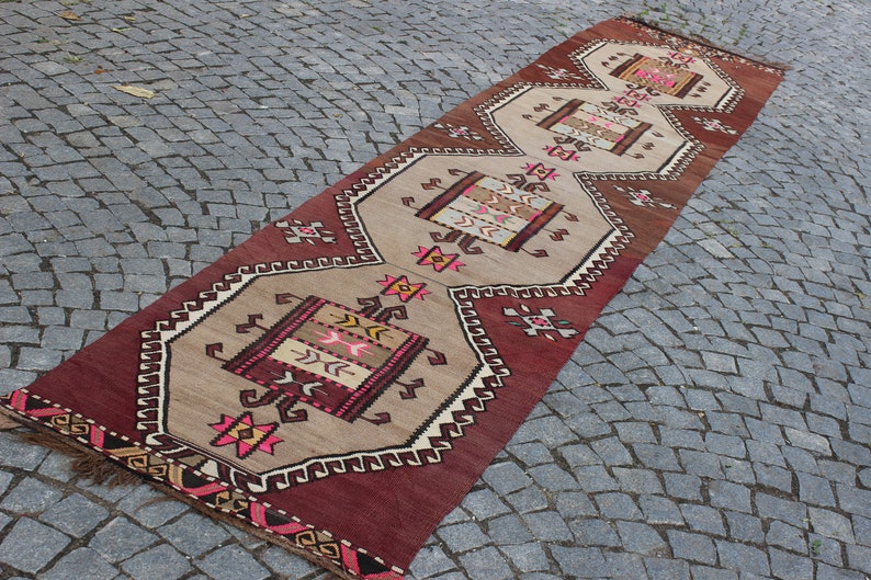 May include: A long, narrow rug with a geometric pattern in shades of red, brown, beige, and pink. The rug has a fringe on both ends.