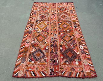 May include: A rectangular textile wall hanging featuring a vibrant patchwork of red, orange, and brown tones. The design includes symmetrical diamond-shaped motifs, intricate geometric embroidery, and striped borders with a fringed bottom edge.