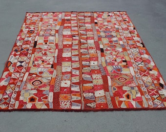 May include: A rectangular, hand-woven rug with a vibrant, geometric design. The rug features a predominantly red background with a patchwork of colorful squares and patterns in shades of orange, white, and blue. The rug's dimensions are not visible.