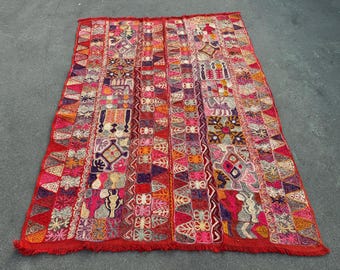 May include: A rectangular textile rug featuring a vibrant red base color with intricate multi-colored embroidery. The design consists of vertical panels filled with geometric shapes, floral motifs, and stylized animal figures, finished with fringe.