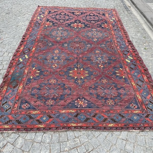 Puede incluir: Una alfombra rectangular grande con un patrón geométrico detallado. La alfombra presenta un fondo rojo oscuro con diseños florales y geométricos azules y amarillos. El borde es rojo más oscuro con detalles azules y amarillos. La alfombra está colocada sobre una superficie de adoquines.