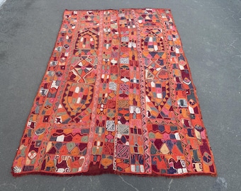 May include: A rectangular, hand-woven rug with a vibrant, geometric design. The rug features a predominantly orange background with a patchwork of colorful shapes, including squares, triangles, and stylized figures. The edges are finished with a fringe.