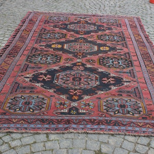 May include: A vintage Turkish rug with a red background and intricate geometric patterns in shades of blue, black, and yellow. The rug has a fringed border.