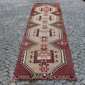 May include: A long, narrow rug with a traditional Turkish design. The rug is beige with red, pink, brown, and yellow geometric patterns. The rug has a fringe on both ends.