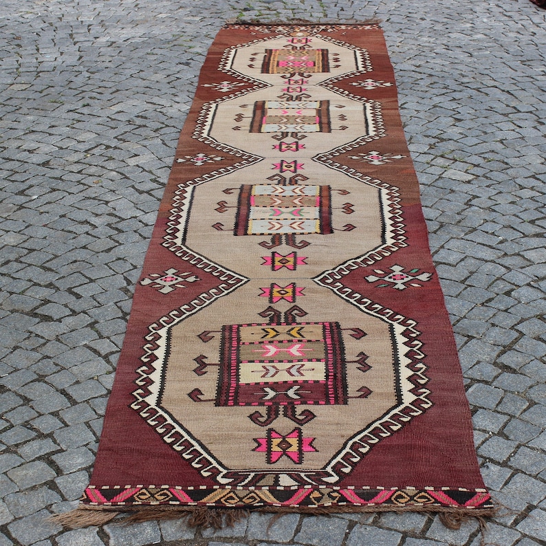 May include: A long, narrow rug with a traditional Turkish design. The rug is beige with red, pink, brown, and yellow geometric patterns. The rug has a fringe on both ends.
