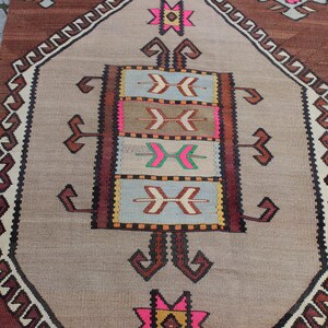 May include: A vintage Turkish rug with a beige background and a central medallion featuring a geometric pattern in shades of brown, pink, and blue. The rug has a burgundy border with a repeating pattern.