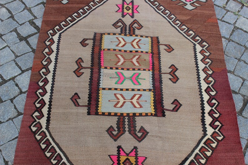 May include: A vintage Turkish rug with a beige background and a central medallion featuring a geometric pattern in shades of brown, pink, and blue. The rug has a burgundy border with a repeating pattern.