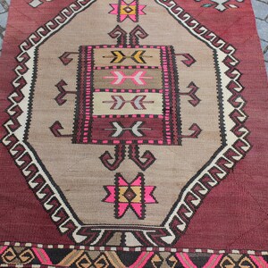 May include: A vintage Turkish rug with a geometric pattern in shades of pink, brown, and white. The rug has a burgundy border with a geometric pattern.