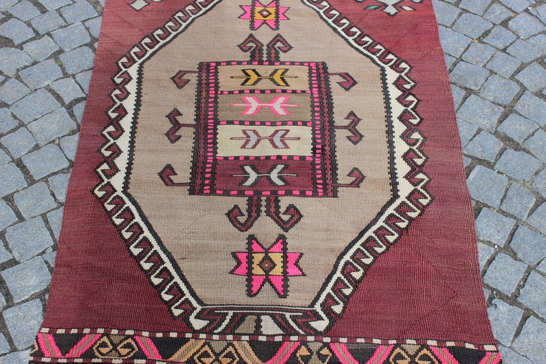 May include: A vintage Turkish rug with a geometric pattern in shades of pink, brown, and white. The rug has a burgundy border with a geometric pattern.