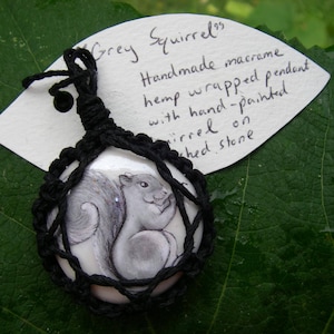 May include: A grey squirrel macrame pendant with a hand-painted image of a squirrel on a white stone. The pendant is made with hemp cord and is wrapped in macrame.