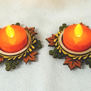 Op de afbeelding: Twee decoratieve kaarshouders met oranje LED-kaarsen. De kaarsen staan in een houten voet omgeven door een bladerontwerp in bruin-, groen- en geeltinten. De kaarshouders zijn ontworpen om op herfstbladeren te lijken.