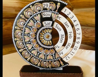 May include: A decorative wooden perpetual calendar featuring a black, gold, and pink mandala design. The calendar shows the date, day, and month, with numbers 1-12 on a curved panel. The base is a dark brown rectangle.