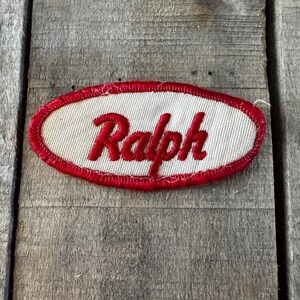 May include: An oval-shaped embroidered patch featuring a white fabric center with a red stitched border. The name Ralph is embroidered in red cursive script across the center of the patch, which has a textured finish and visible thread details.