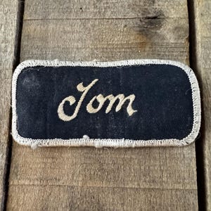 May include: A rectangular black fabric patch with rounded corners and a white stitched border. The word Jom is embroidered in the center in a light beige cursive script. The patch features a textured surface and visible thread details throughout.
