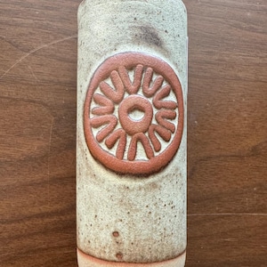 May include: A white ceramic vase with a brown, circular design. The design features a stylized sun with a circle in the center.