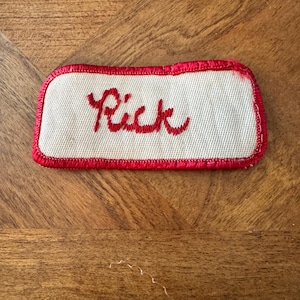 May include: A rectangular embroidered patch with a white fabric base and a red stitched border. The name Rick is embroidered in red cursive script in the center of the patch. The patch has rounded corners and a textured, woven appearance.