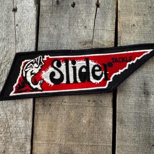 May include: A trapezoidal embroidered patch featuring a black border and a red center. The patch displays a white graphic of a fish jumping and the black text Slider Tackle. The design is shaped like the state of Tennessee with a black background.