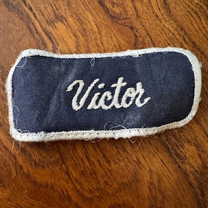 May include: A rectangular navy blue fabric patch with rounded corners and a white stitched border. The name Victor is embroidered in white cursive script across the center of the patch. The fabric has a visible weave and some loose threads.