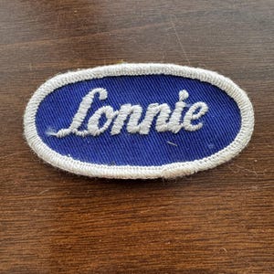 Puede incluir: Parche ovalado con el nombre "Lonnie" bordado en blanco sobre fondo azul. El parche tiene un borde blanco y probablemente sea para un uniforme o prenda de vestir.