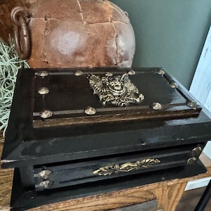 May include: A rectangular black wooden box featuring a metallic gold-toned crest of a double-headed eagle on the lid. The box is accented with decorative gold-toned metal studs along the edges and a horizontal gold-toned handle on the front drawer.