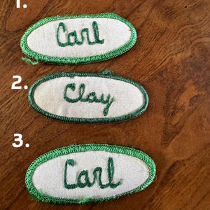 May include: Three vintage name patches. Two patches read "Carl" in green script, and one reads "Clay". Each patch has a white oval center with a green stitched border. The patches are on a wooden surface.