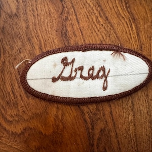 May include: An oval-shaped fabric patch with a white background and a dark brown embroidered border. The name Greg is stitched in the center in dark brown cursive script. A thin, horizontal dark brown line runs across the white fabric.