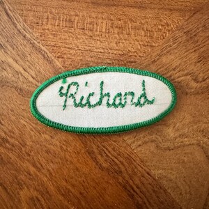 Puede incluir: Un parche de tela ovalado con fondo blanco y borde bordado en verde. El nombre Richard está cosido en el centro con hilo verde en letra cursiva. El parche presenta un borde limpio y acabado con un denso bordado de color verde.